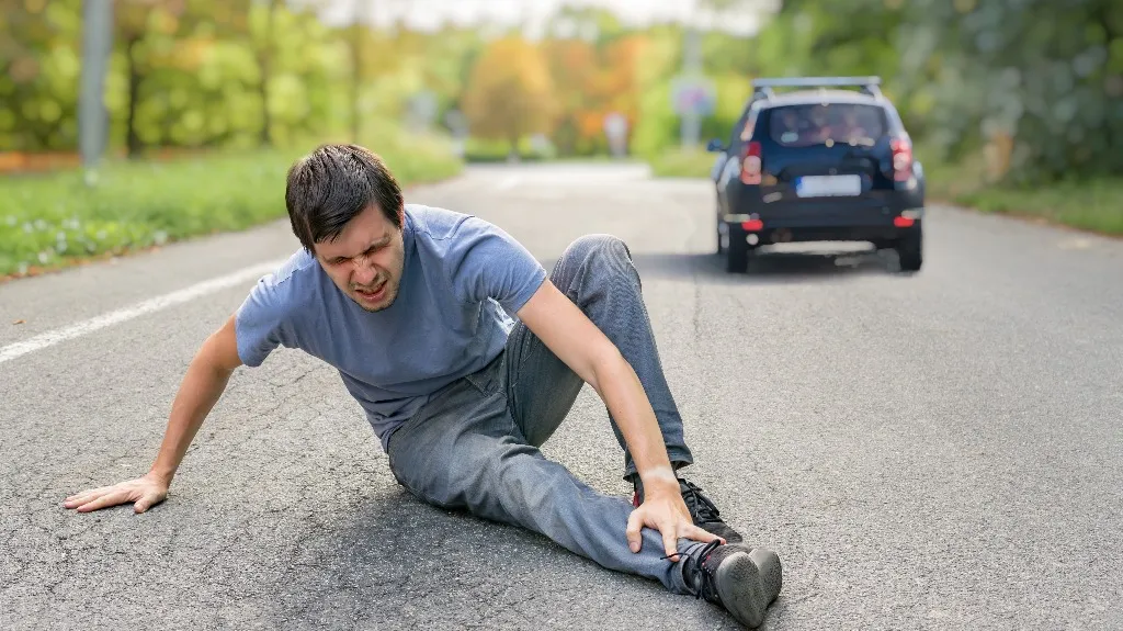 Man in blue shirt and gray pants sitting on road, holding injured leg with car in background.