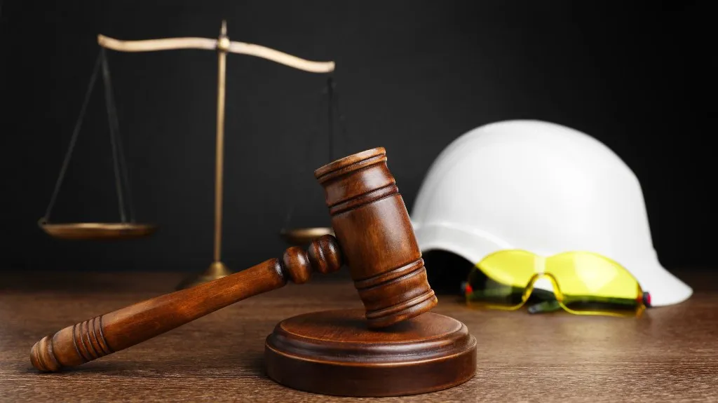 Wooden judge's gavel on table with brass scales, white hard hat, and yellow safety glasses.