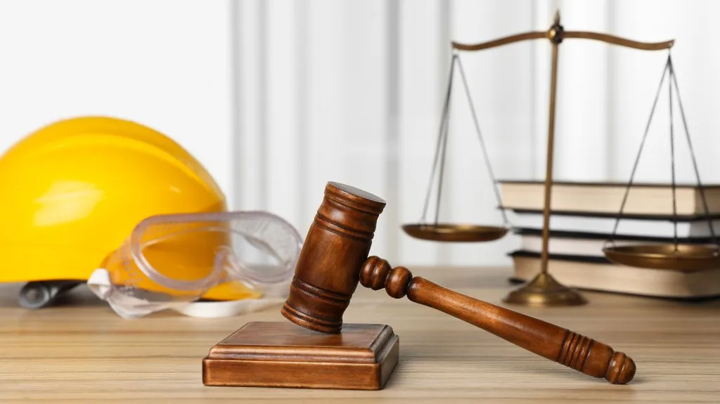 Wooden judge gavel, yellow construction helmet, safety goggles, scales of justice, and stacked books on wooden surface.