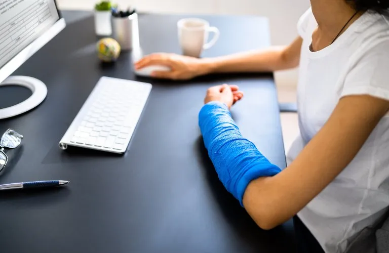 Person with blue arm cast sitting at black desk with computer keyboard, pen, and coffee mug.