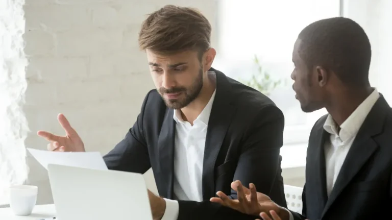 Two professionally dressed men engaged in a discussion, one holding a document, seated at a desk with a laptop.