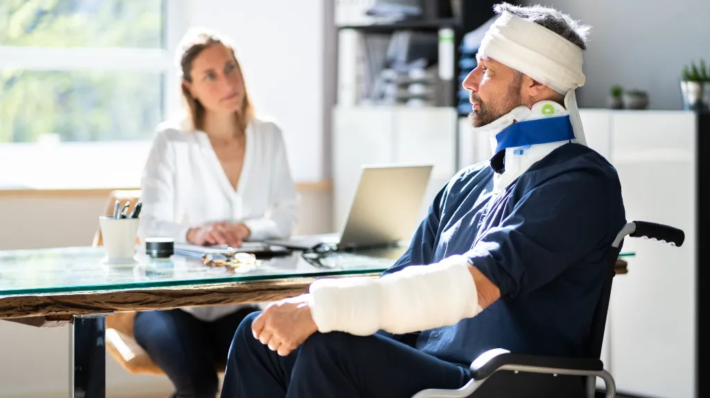 Man with head, neck, and arm injuries seated in wheelchair, woman in white shirt at desk.