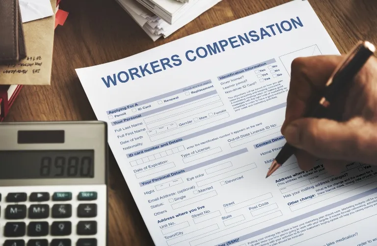 Worker filling out a workers compensation form on a wooden desk with a calculator nearby.