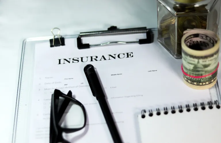 Insurance form on clipboard with pen, eyeglasses, rolled cash, and jar of coins.